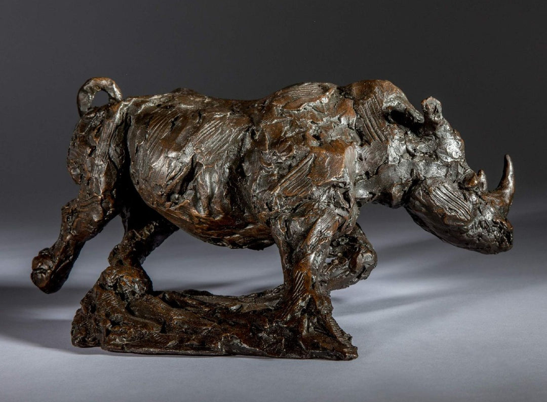 Bronze sculpture of a white rhino mid-charge, captured with bold textures and muscular form, evoking movement and strength.