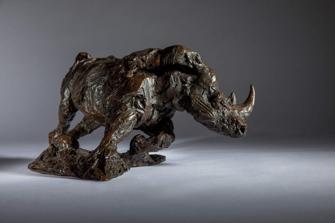 Bronze sculpture of a white rhino mid-charge, captured with bold textures and muscular form, evoking movement and strength.