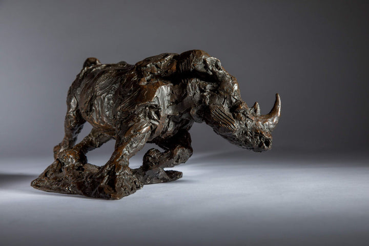 Bronze sculpture of a white rhino mid-charge, captured with bold textures and muscular form, evoking movement and strength.