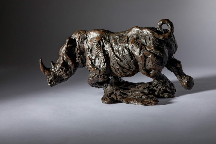 Bronze sculpture of a white rhino mid-charge, captured with bold textures and muscular form, evoking movement and strength.