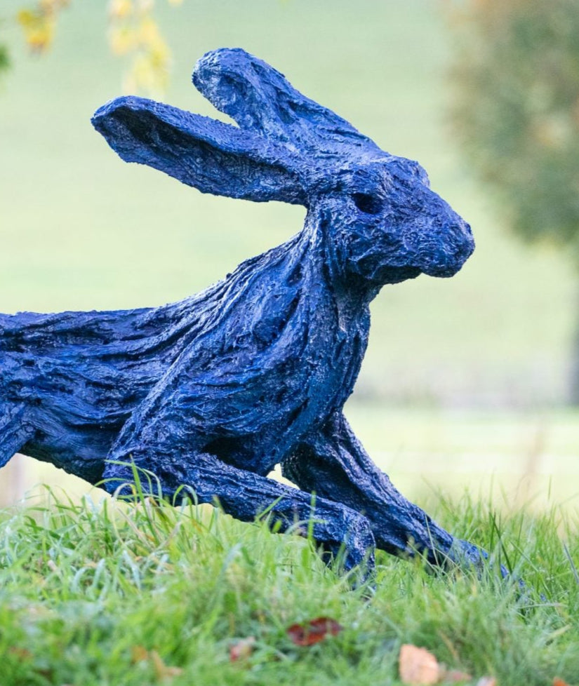 Large bronze sculpture of a hare mid-sprint, finished in a striking electric blue patina with exaggerated limbs and twisting motion, set outdoors in a lush grassy landscape with autumn trees in the background.