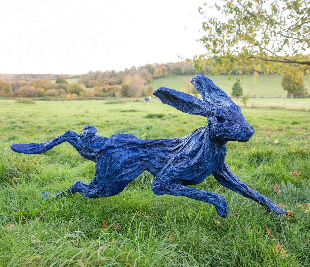Large bronze sculpture of a hare mid-sprint, finished in a striking electric blue patina with exaggerated limbs and twisting motion, set outdoors in a lush grassy landscape with autumn trees in the background.