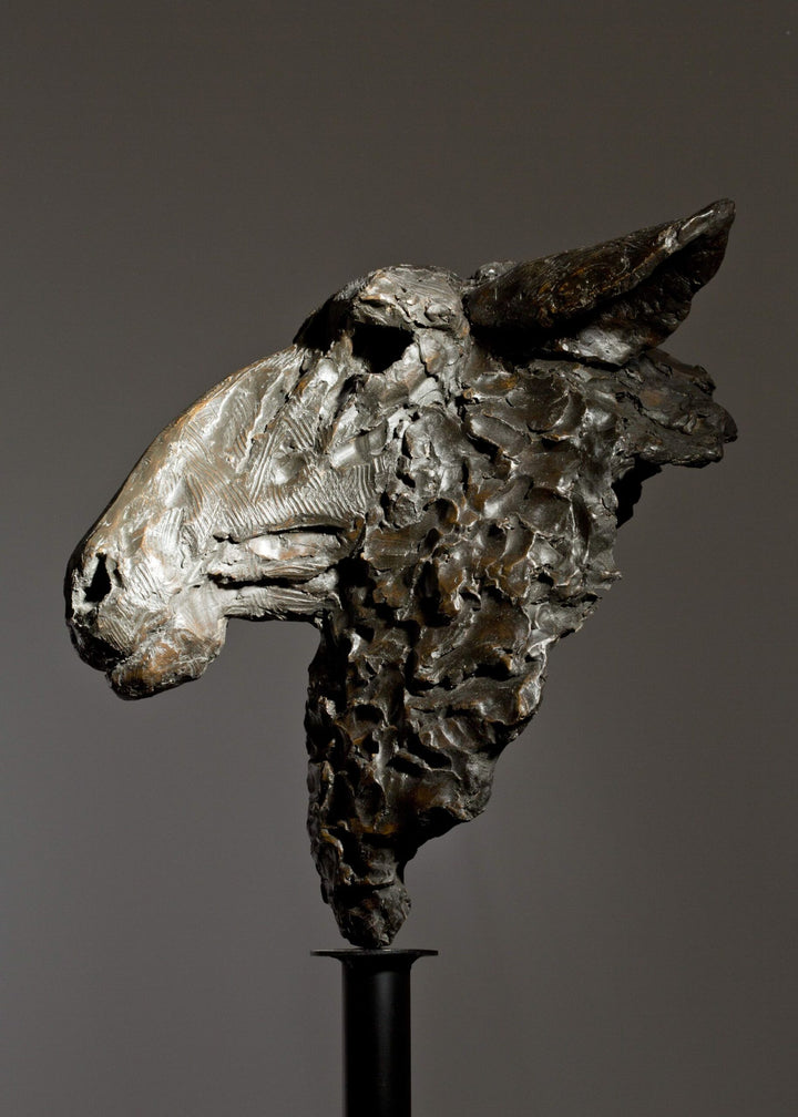 Life-size bronze sculpture of a sheep’s head with textured fleece and alert, characterful expression, mounted on a tall dark metal stand.