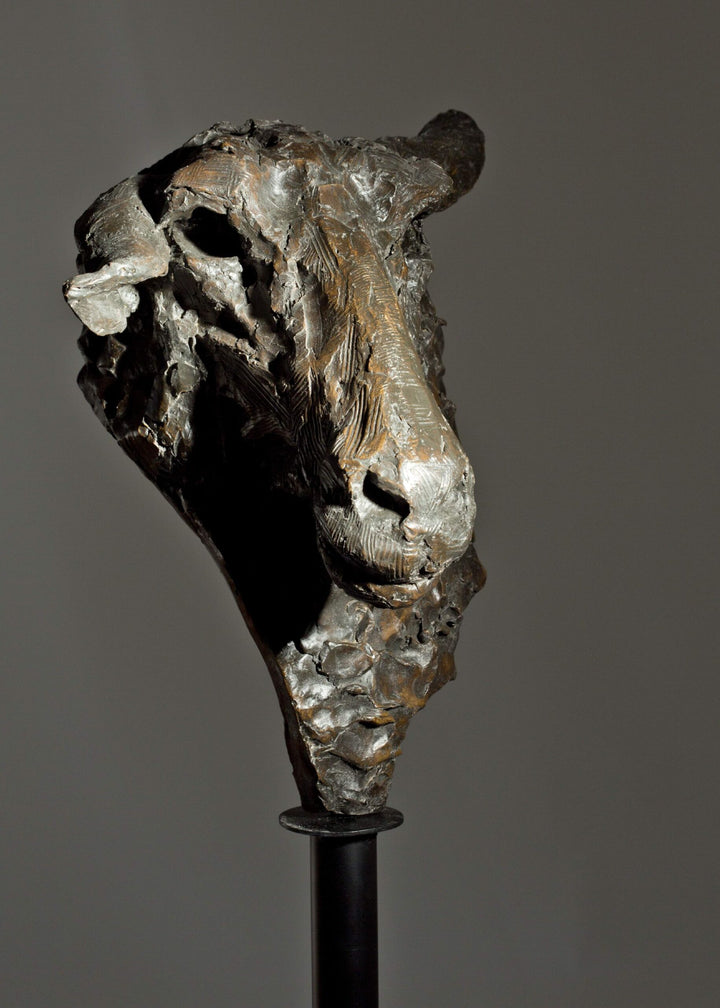 Life-size bronze sculpture of a sheep’s head with textured fleece and alert, characterful expression, mounted on a tall dark metal stand.