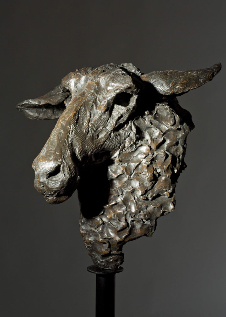 Life-size bronze sculpture of a sheep’s head with textured fleece and alert, characterful expression, mounted on a tall dark metal stand.