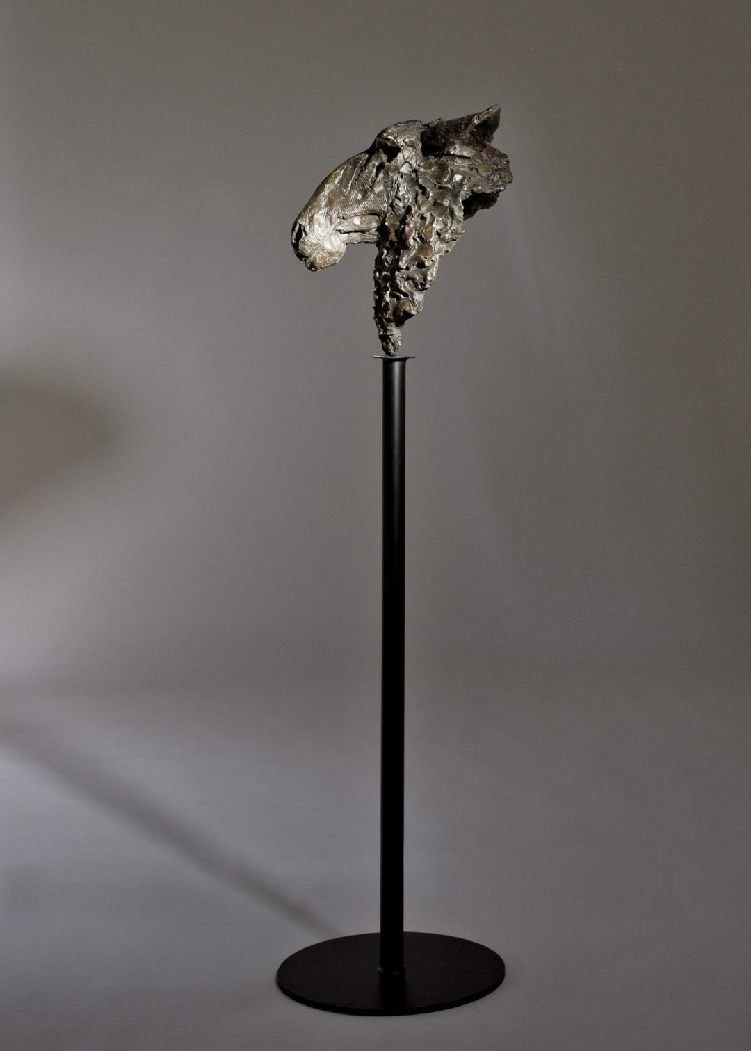 Life-size bronze sculpture of a sheep’s head with textured fleece and alert, characterful expression, mounted on a tall dark metal stand.