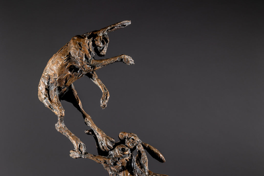 Bronze sculpture of two dynamic boxing hares mid-air, captured in a lively and expressive pose with interlinked limbs and alert ears, mounted on a sleek dark base and presented against a neutral backdrop.
