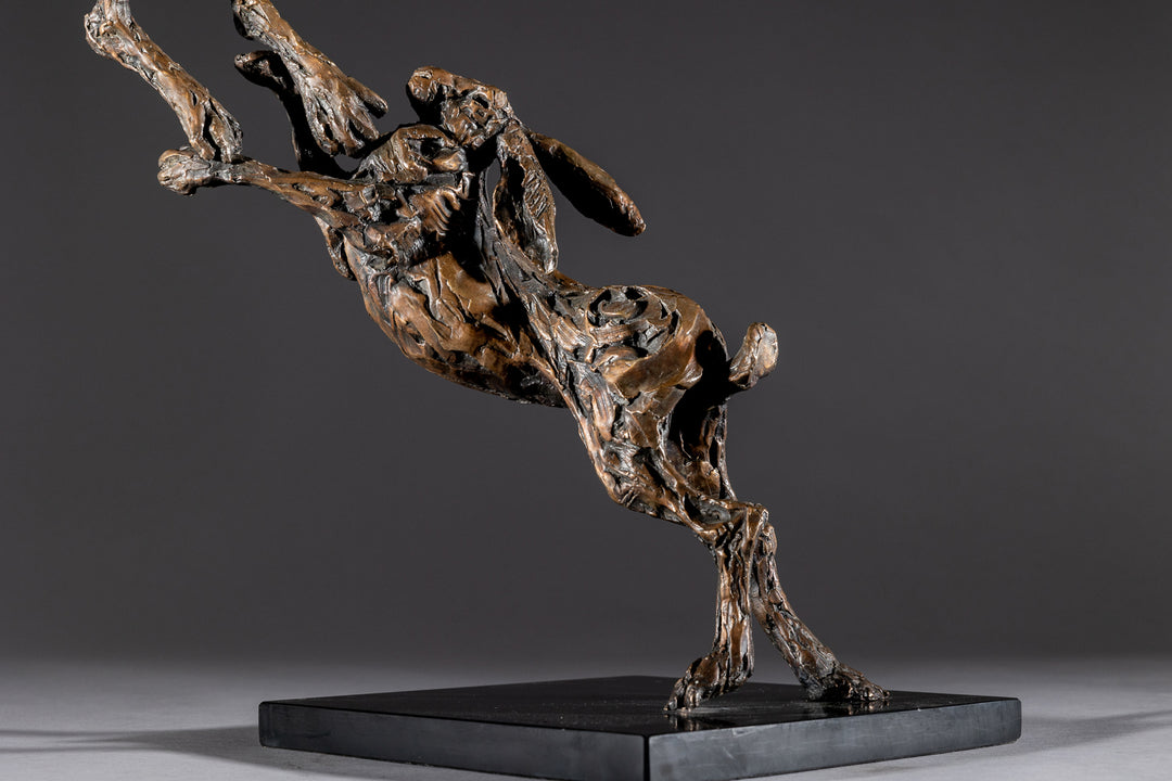 Bronze sculpture of two dynamic boxing hares mid-air, captured in a lively and expressive pose with interlinked limbs and alert ears, mounted on a sleek dark base and presented against a neutral backdrop.