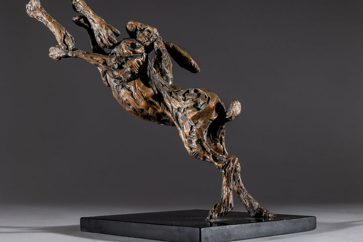 Bronze sculpture of two dynamic boxing hares mid-air, captured in a lively and expressive pose with interlinked limbs and alert ears, mounted on a sleek dark base and presented against a neutral backdrop.
