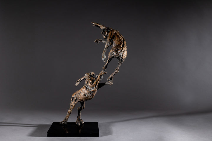 Bronze sculpture of two dynamic boxing hares mid-air, captured in a lively and expressive pose with interlinked limbs and alert ears, mounted on a sleek dark base and presented against a neutral backdrop.