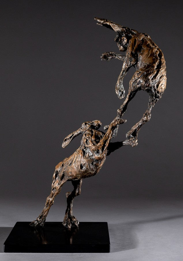 Bronze sculpture of two dynamic boxing hares mid-air, captured in a lively and expressive pose with interlinked limbs and alert ears, mounted on a sleek dark base and presented against a neutral backdrop.