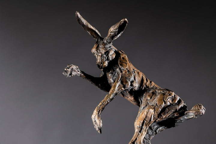 Bronze sculpture of two dynamic boxing hares mid-air, captured in a lively and expressive pose with interlinked limbs and alert ears, mounted on a sleek dark base and presented against a neutral backdrop.