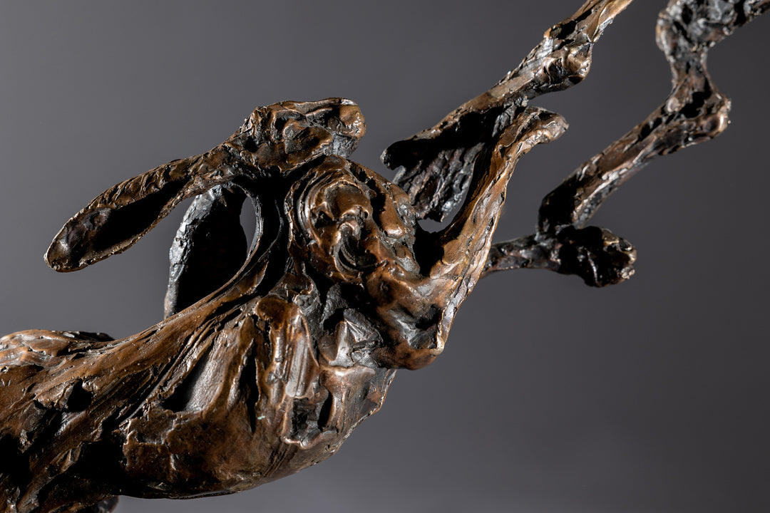 Bronze sculpture of two dynamic boxing hares mid-air, captured in a lively and expressive pose with interlinked limbs and alert ears, mounted on a sleek dark base and presented against a neutral backdrop.
