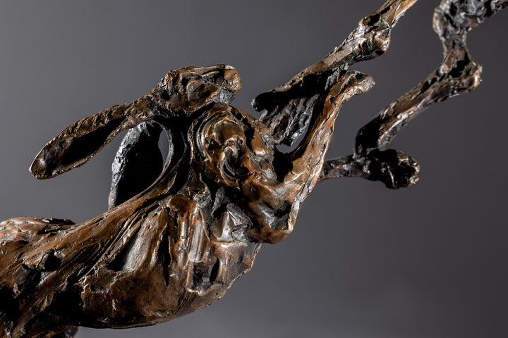 Bronze sculpture of two dynamic boxing hares mid-air, captured in a lively and expressive pose with interlinked limbs and alert ears, mounted on a sleek dark base and presented against a neutral backdrop.