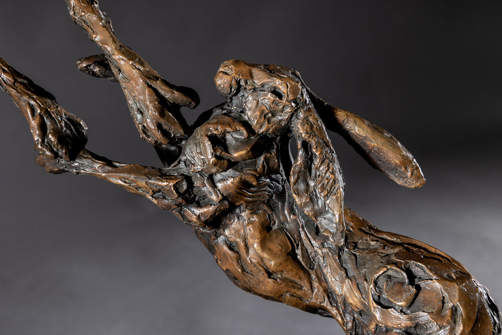 Bronze sculpture of two dynamic boxing hares mid-air, captured in a lively and expressive pose with interlinked limbs and alert ears, mounted on a sleek dark base and presented against a neutral backdrop.