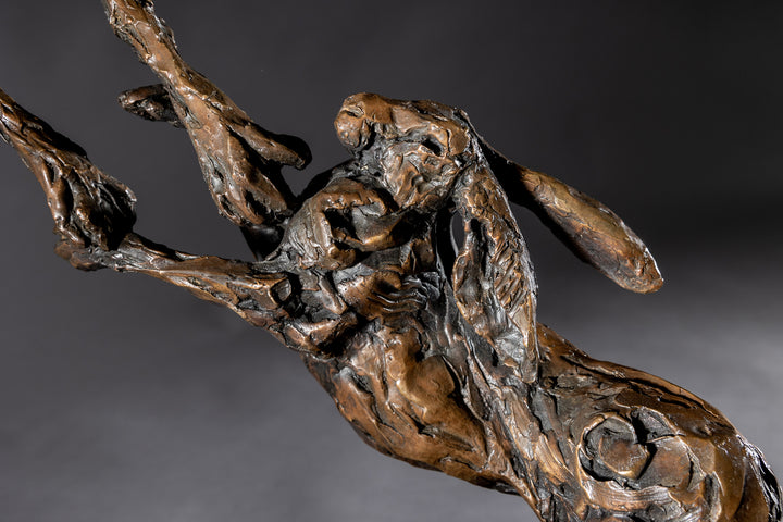 Bronze sculpture of two dynamic boxing hares mid-air, captured in a lively and expressive pose with interlinked limbs and alert ears, mounted on a sleek dark base and presented against a neutral backdrop.