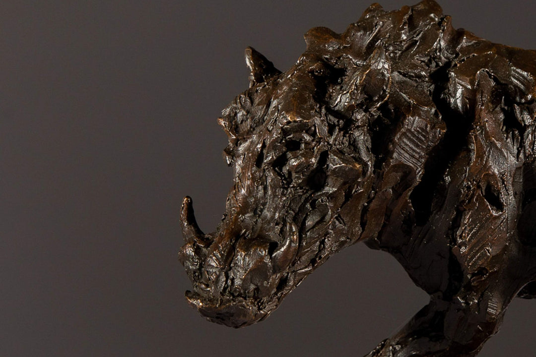 Bronze sculpture of a warthog mid-sprint with exaggerated texture and raised tail, captured in dynamic motion and set against a neutral backdrop.