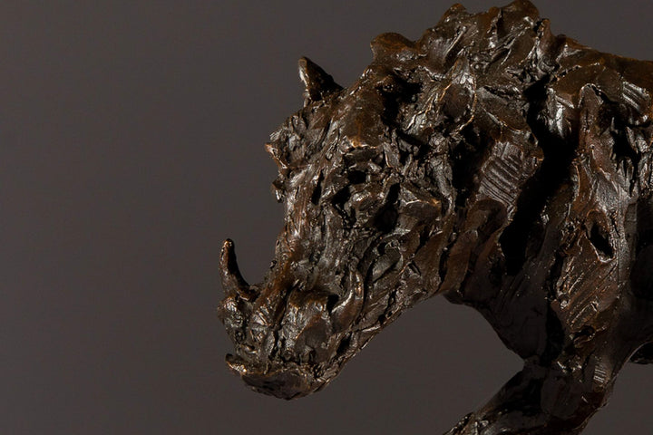Bronze sculpture of a warthog mid-sprint with exaggerated texture and raised tail, captured in dynamic motion and set against a neutral backdrop.