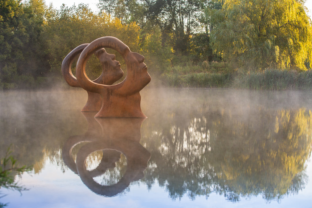 Simon Gudgeon bronze sculpture Search for Enlightenment featuring monumental interlocking abstract human forms with circular openings displayed within a landscaped outdoor setting.