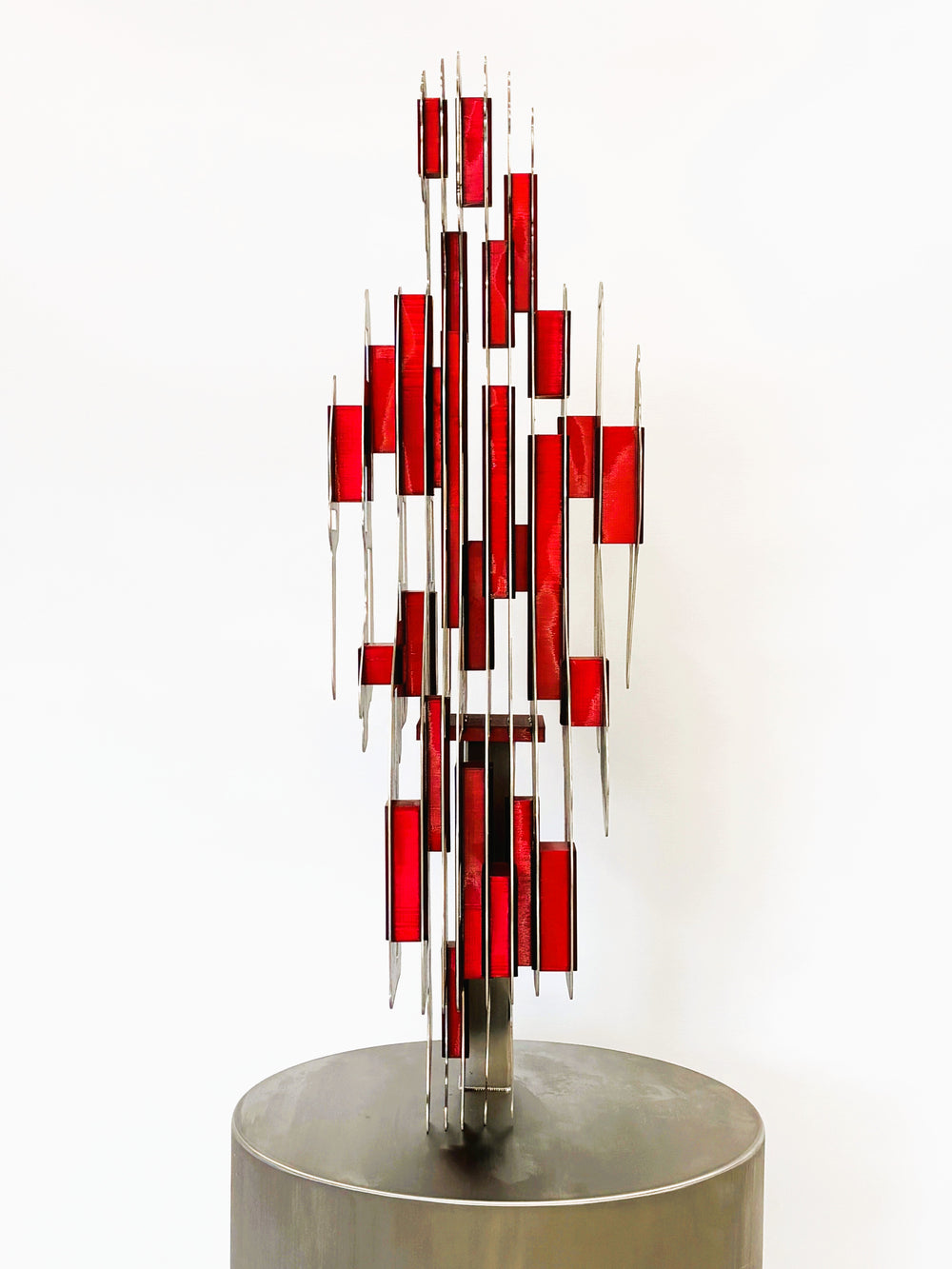 Sculpture titled Seated Girl I by Tom Hiscox, composed of layered stainless steel with red resin inserts forming a stylised seated female figure, shown from multiple angles on a brushed circular base.