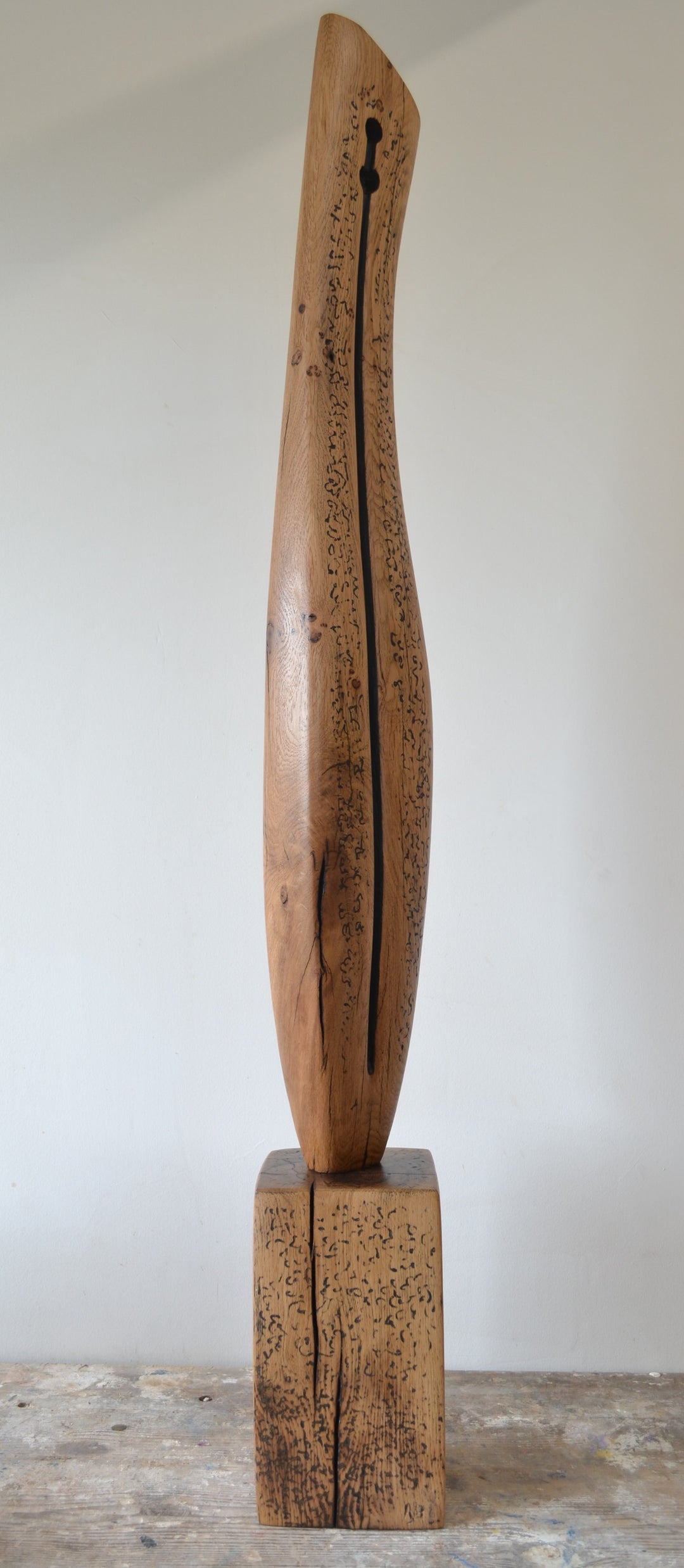 Sentinel by Nicola Beattie – tall, hand-carved oak sculpture with a smooth tapering profile and deep central black inlay ending in a keyhole opening. Warm beige oak with blackened markings and natural cracks, echoing ancient standing stones in a minimalist, monumental style.