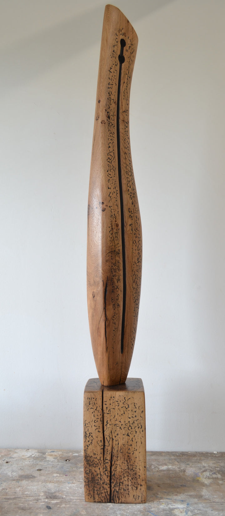 Sentinel by Nicola Beattie – tall, hand-carved oak sculpture with a smooth tapering profile and deep central black inlay ending in a keyhole opening. Warm beige oak with blackened markings and natural cracks, echoing ancient standing stones in a minimalist, monumental style.