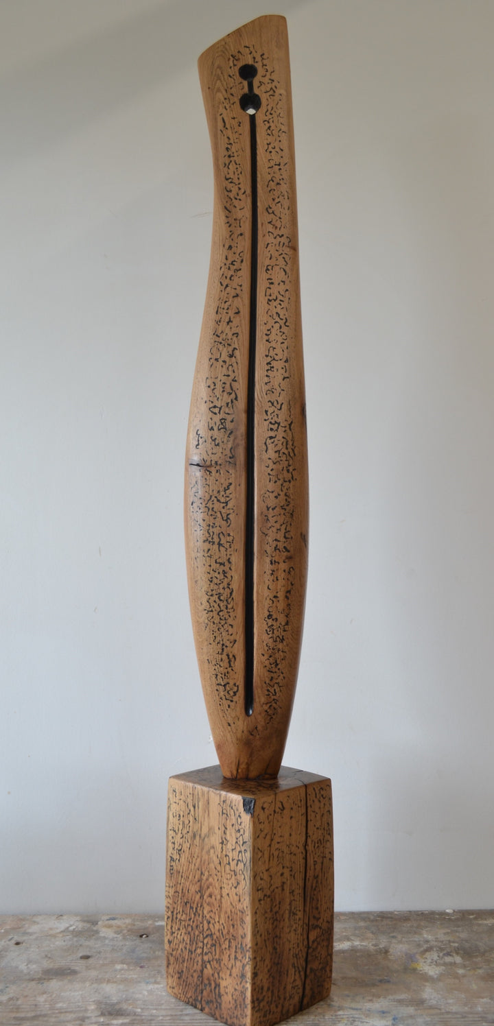 Sentinel by Nicola Beattie – tall, hand-carved oak sculpture with a smooth tapering profile and deep central black inlay ending in a keyhole opening. Warm beige oak with blackened markings and natural cracks, echoing ancient standing stones in a minimalist, monumental style.