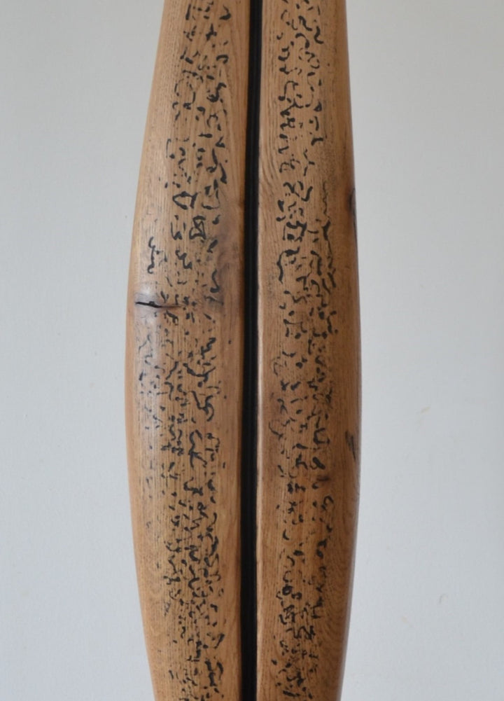 Sentinel by Nicola Beattie – tall, hand-carved oak sculpture with a smooth tapering profile and deep central black inlay ending in a keyhole opening. Warm beige oak with blackened markings and natural cracks, echoing ancient standing stones in a minimalist, monumental style.