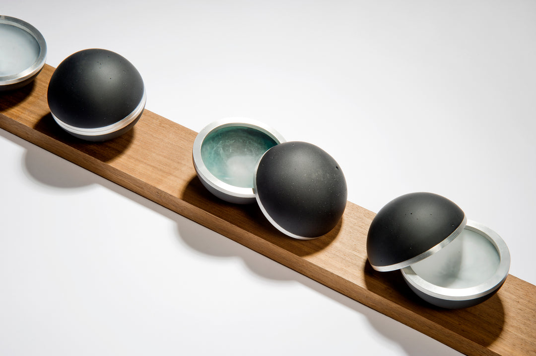 Serendipity by Tracy Nicholls — a unique glass sculpture of five black lead crystal spheres resting on a walnut base. Each sphere is formed from four lost wax cast hemispheres, the opaque black outer shells contrasting with luminous interiors of layered coloured glass. Aluminium rings and concealed neodymium magnets connect the hemispheres, allowing them to open and reveal swirling patterns of colour created by chance during the casting process. The polished surfaces and precise engineering contrast with th