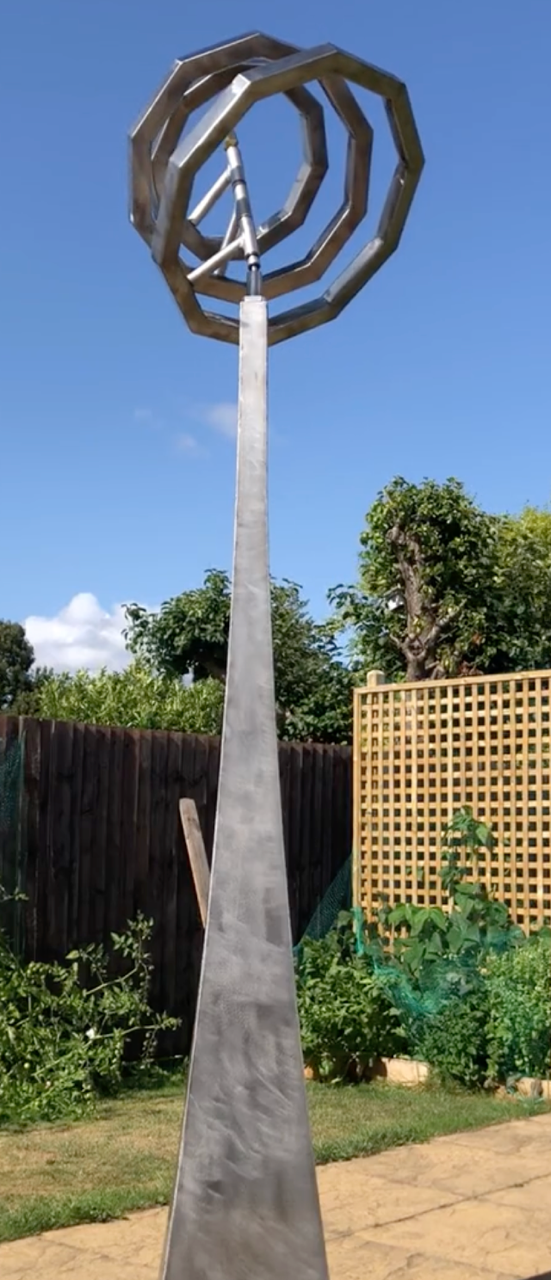 Martin Jarvis kinetic sculpture titled Serenity, featuring interlocking octagonal rings crafted from polished stainless steel. The rings are mounted atop a tall, tapered plinth and rotate gently in the breeze, creating a mesmerising visual rhythm that contrasts geometric precision with organic movement.