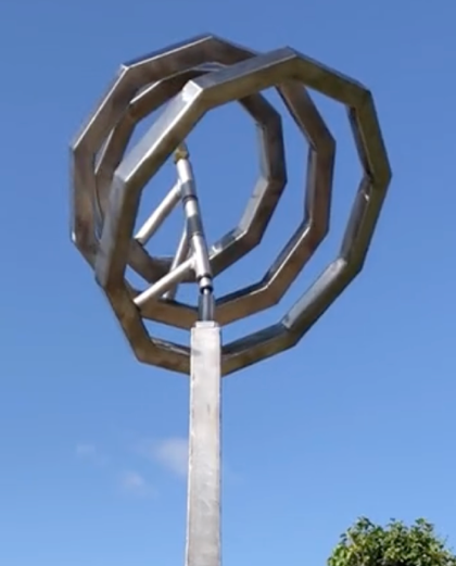 Martin Jarvis kinetic sculpture titled Serenity, featuring interlocking octagonal rings crafted from polished stainless steel. The rings are mounted atop a tall, tapered plinth and rotate gently in the breeze, creating a mesmerising visual rhythm that contrasts geometric precision with organic movement.