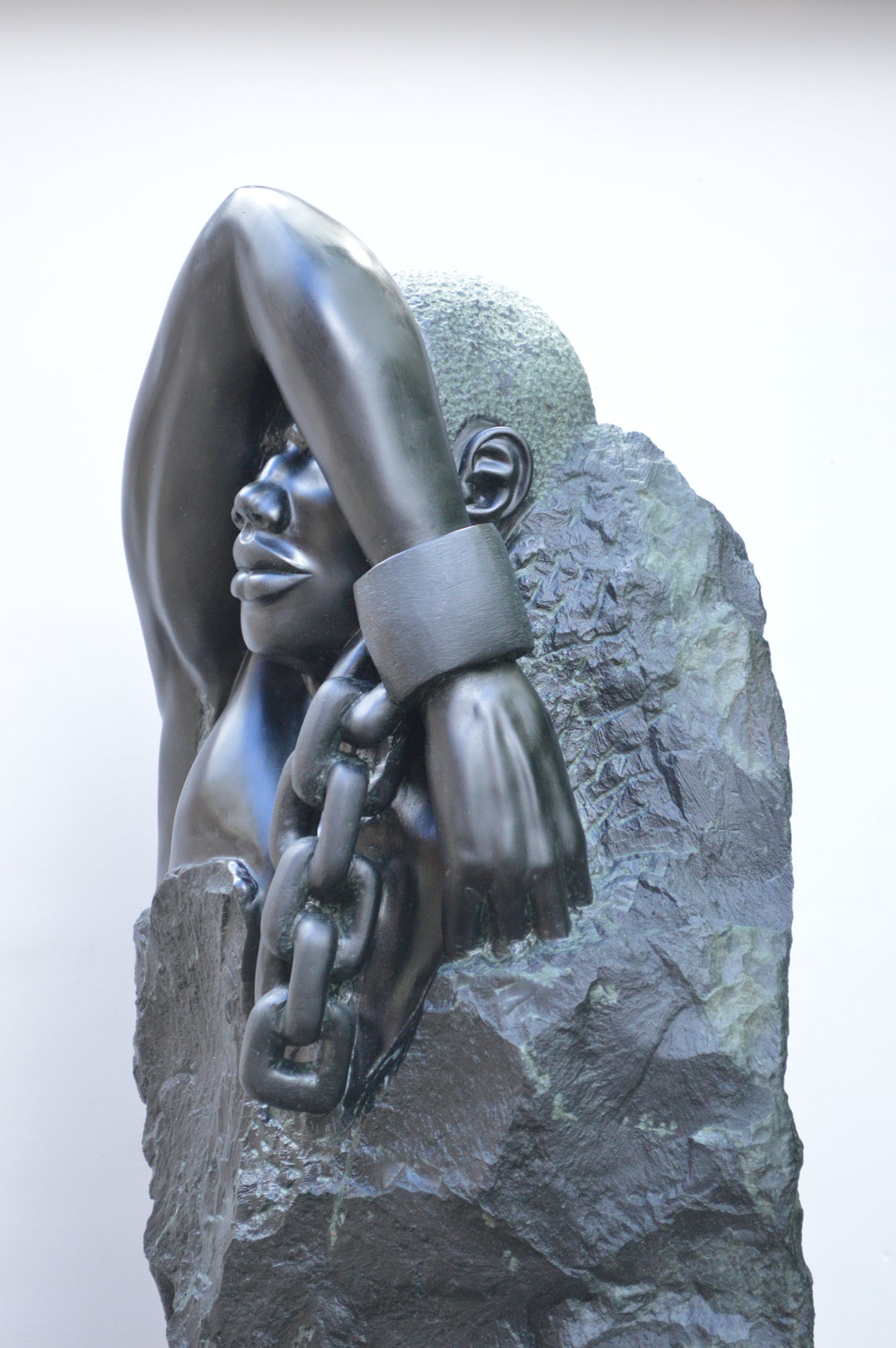 Deborah Harrison sculpture Slave Song, a one-off hand-carved black soapstone figure showing a bound male form with shackled wrist and heavy chain links, emerging from raw stone. The polished dark surface contrasts against the rough, natural texture, creating a powerful narrative of struggle, endurance, and the human spirit.