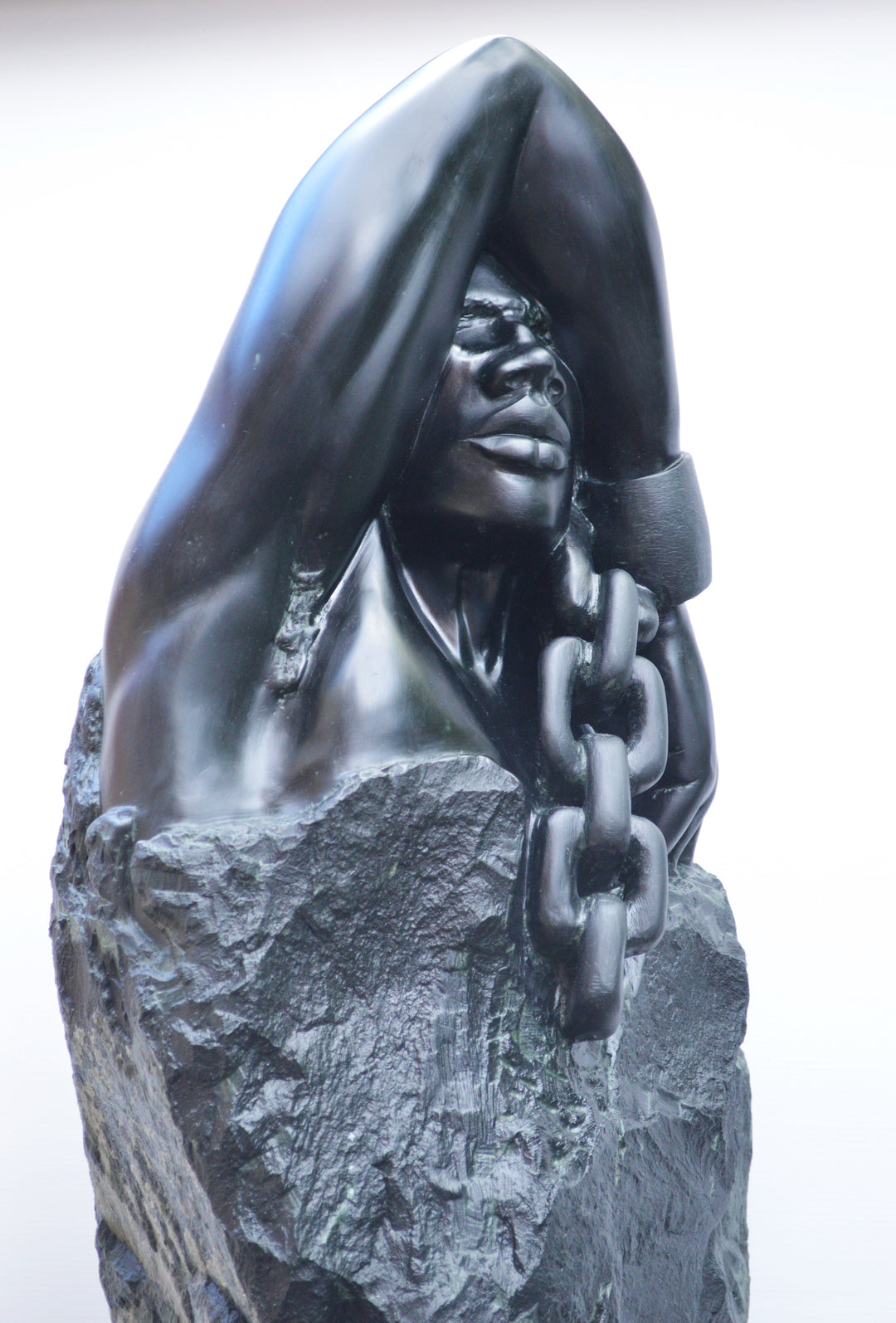 Deborah Harrison sculpture Slave Song, a one-off hand-carved black soapstone figure showing a bound male form with shackled wrist and heavy chain links, emerging from raw stone. The polished dark surface contrasts against the rough, natural texture, creating a powerful narrative of struggle, endurance, and the human spirit.