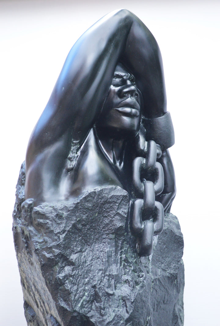 Deborah Harrison sculpture Slave Song, a one-off hand-carved black soapstone figure showing a bound male form with shackled wrist and heavy chain links, emerging from raw stone. The polished dark surface contrasts against the rough, natural texture, creating a powerful narrative of struggle, endurance, and the human spirit.