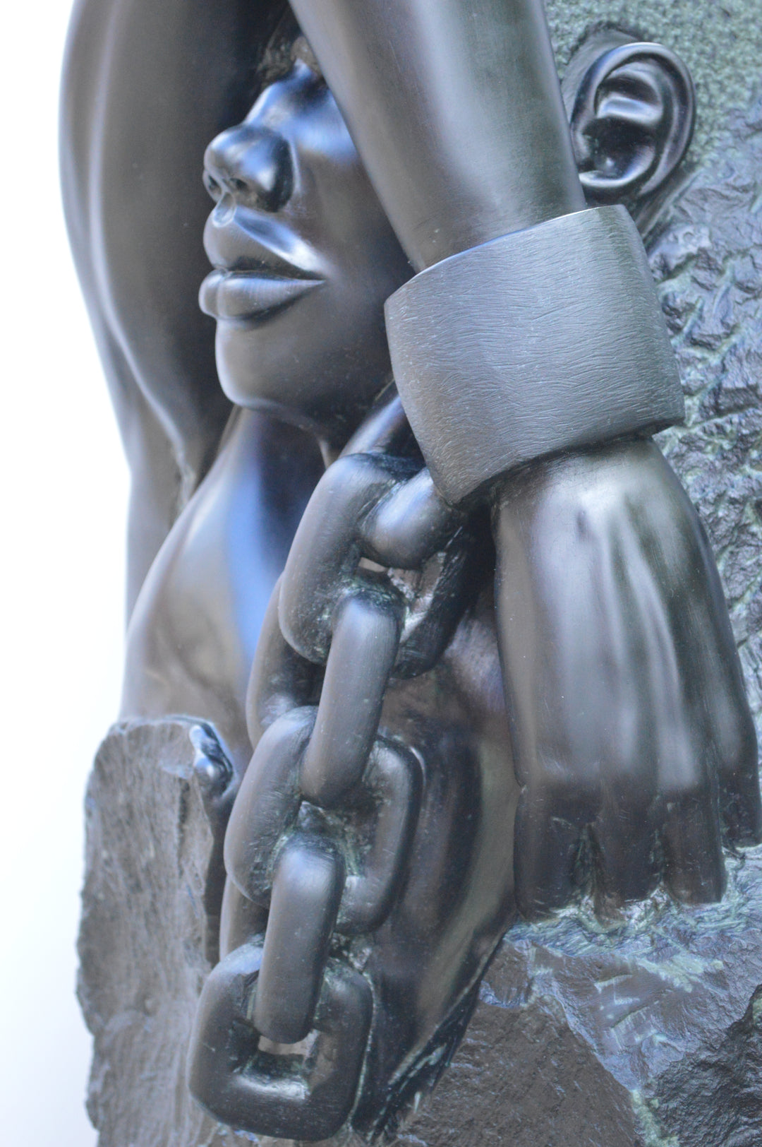 Deborah Harrison sculpture Slave Song, a one-off hand-carved black soapstone figure showing a bound male form with shackled wrist and heavy chain links, emerging from raw stone. The polished dark surface contrasts against the rough, natural texture, creating a powerful narrative of struggle, endurance, and the human spirit.