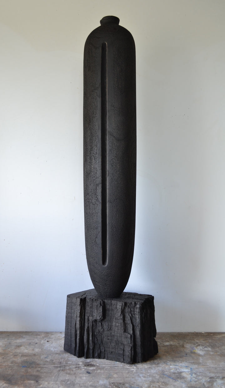 Solitude by Nicola Beattie – tall hand-carved sculpture in charred sycamore and bog oak, with elongated vertical form, deep central recess, and textured blackened surface on rugged base, evoking ancient standing stones and quiet resilience.