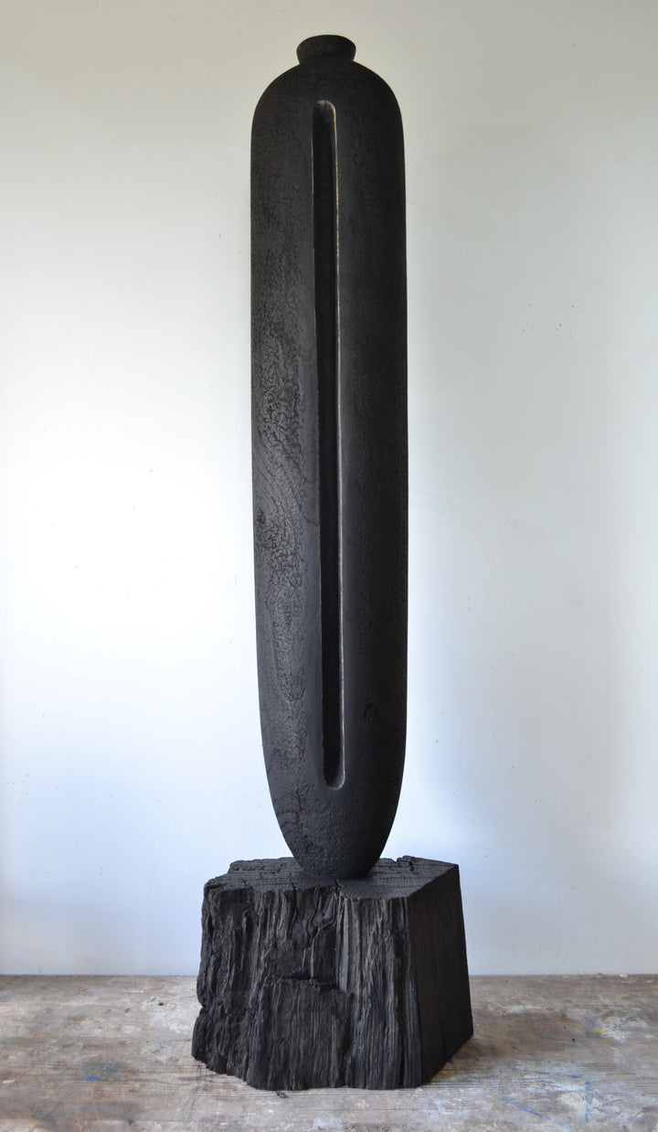 Solitude by Nicola Beattie – tall hand-carved sculpture in charred sycamore and bog oak, with elongated vertical form, deep central recess, and textured blackened surface on rugged base, evoking ancient standing stones and quiet resilience.