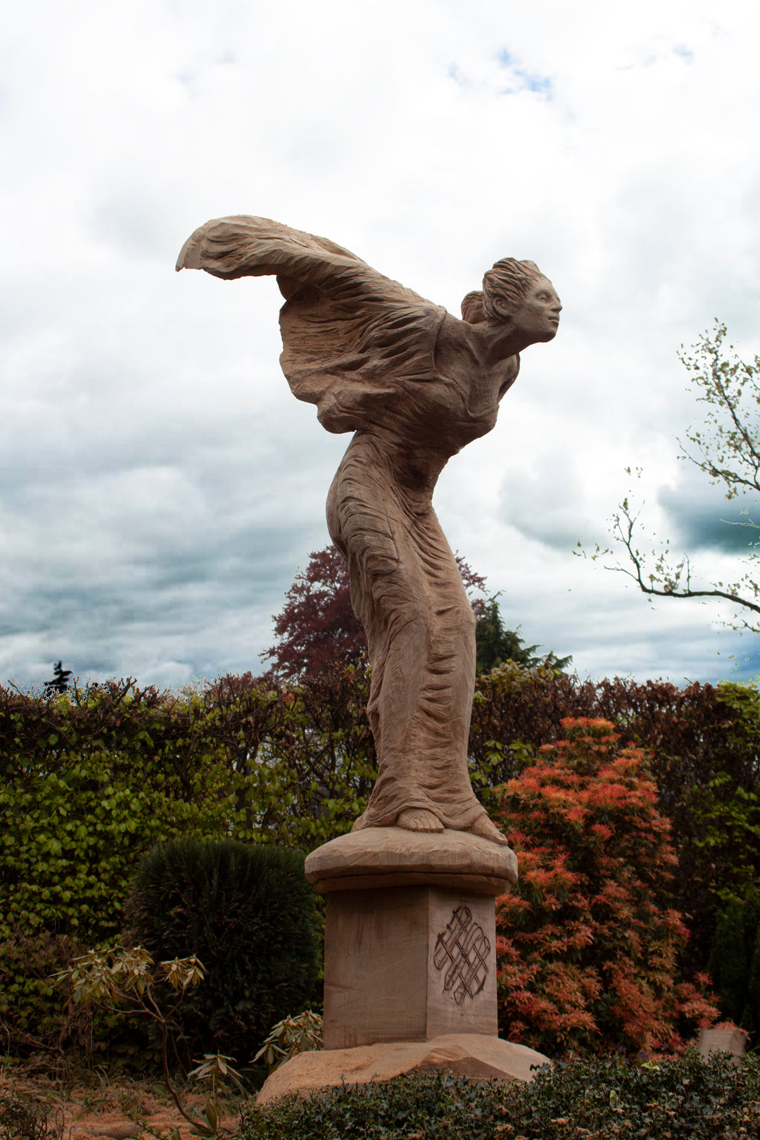 Tall wooden sculpture of a woman in flowing robes leaning forward against the wind, mounted on a pedestal in a landscaped garden. Her arms are swept back and hair tied, conveying a sense of motion and resilience beneath a dramatic cloudy sky.
