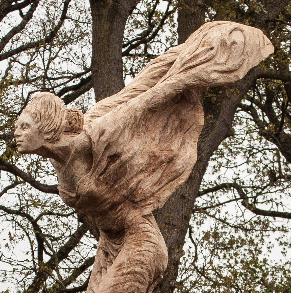 Tall wooden sculpture of a woman in flowing robes leaning forward against the wind, mounted on a pedestal in a landscaped garden. Her arms are swept back and hair tied, conveying a sense of motion and resilience beneath a dramatic cloudy sky.