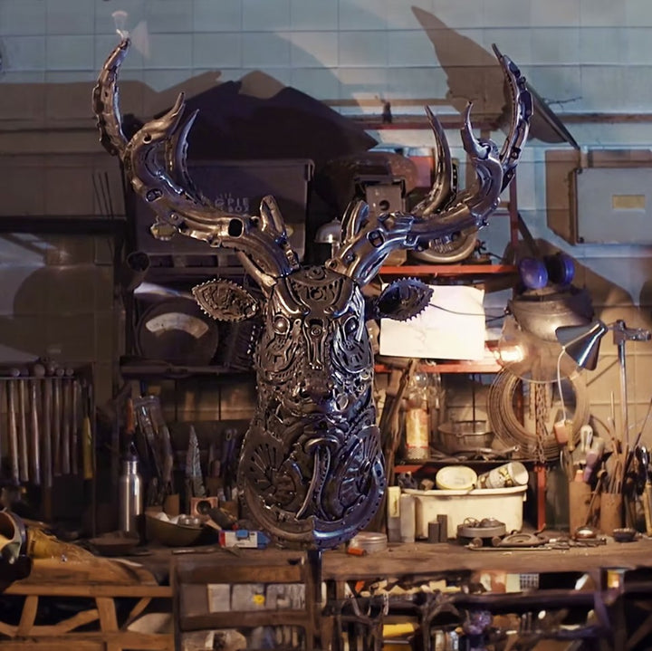 Sculpted stag head made from welded metal parts and gears, mounted against a white background.
