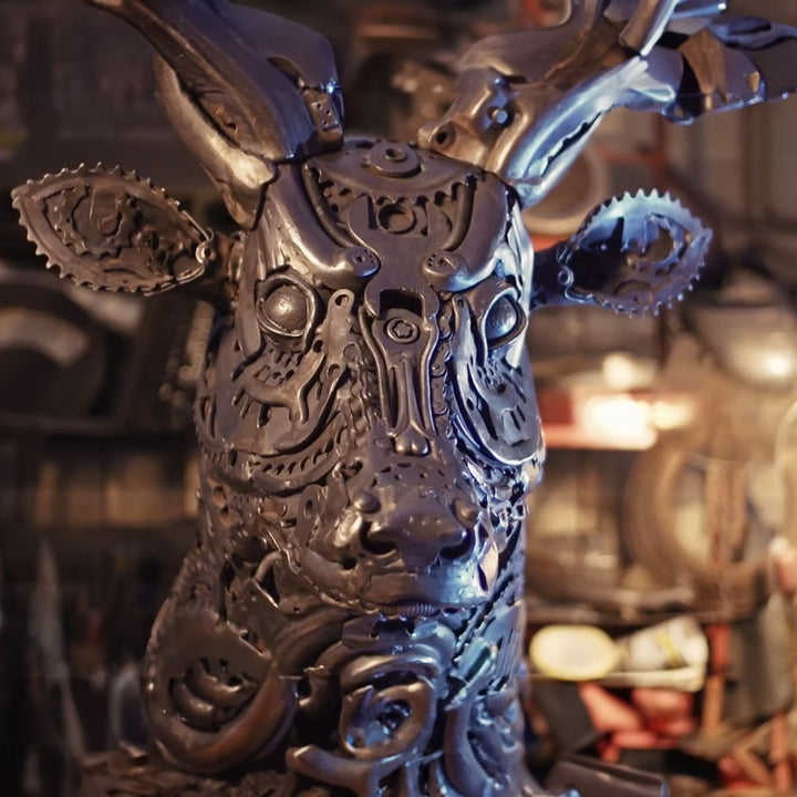 Sculpted stag head made from welded metal parts and gears, mounted against a white background.
