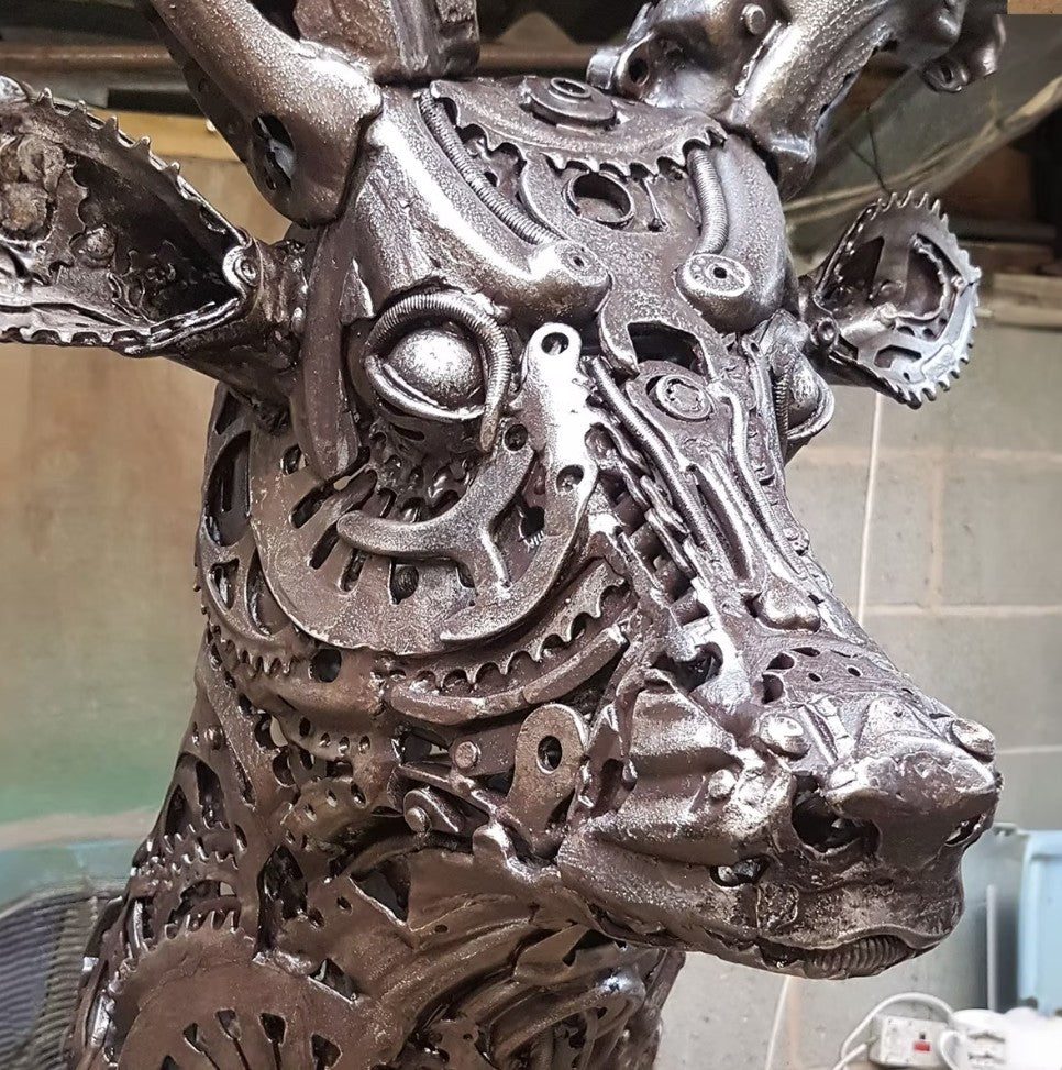 Sculpted stag head made from welded metal parts and gears, mounted against a white background.
