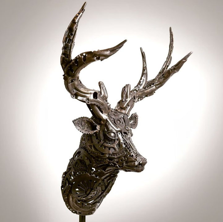 Sculpted stag head made from welded metal parts and gears, mounted against a white background.
