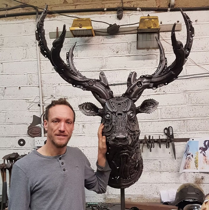 Sculpted stag head made from welded metal parts and gears, mounted against a white background.
