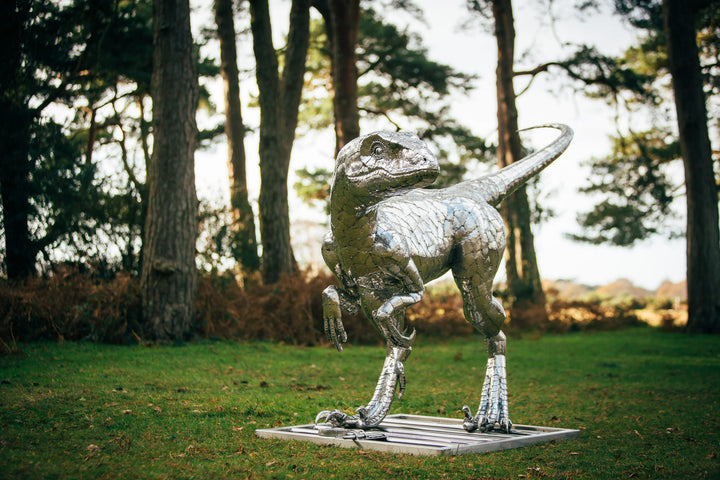 ‘Velociraptor’ by Michael Turner, a unique, one-of-one stainless steel dinosaur sculpture depicting a powerful velociraptor in an alert, dynamic pose. Handcrafted from intricately shaped and welded stainless steel, the sculpture features a polished silver finish, individually detailed metallic scales, muscular hind legs, sharp curved claws, smaller forelimbs and a long, sweeping tail. Standing on a rectangular stainless steel base, this large-scale contemporary sculpture captures the agility and predatory c