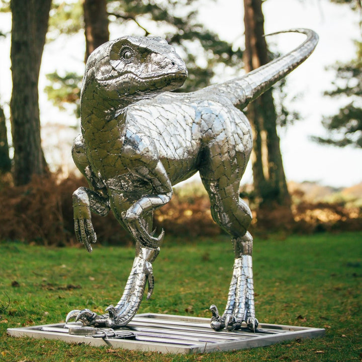 ‘Velociraptor’ by Michael Turner, a unique, one-of-one stainless steel dinosaur sculpture depicting a powerful velociraptor in an alert, dynamic pose. Handcrafted from intricately shaped and welded stainless steel, the sculpture features a polished silver finish, individually detailed metallic scales, muscular hind legs, sharp curved claws, smaller forelimbs and a long, sweeping tail. Standing on a rectangular stainless steel base, this large-scale contemporary sculpture captures the agility and predatory c