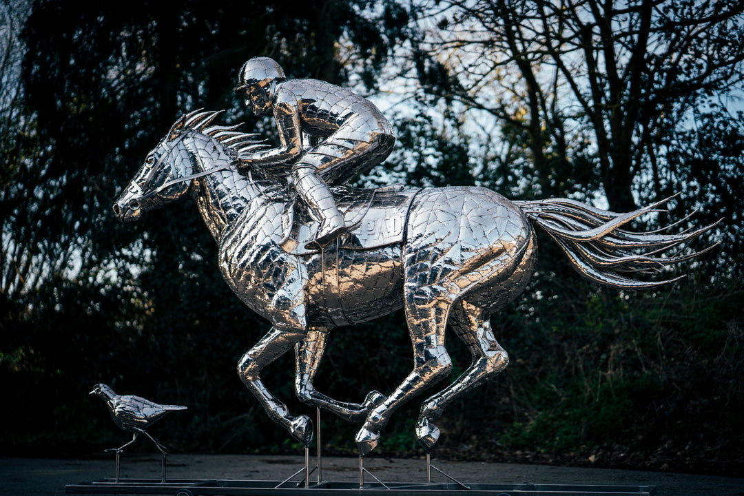 ‘Racehorse & Jockey - A Dynamic Force’ by Michael Turner, a unique, one-of-one stainless steel equestrian sculpture depicting a powerful thoroughbred racehorse galloping at full speed with its jockey leaning forward in a racing position. Handcrafted from hundreds of individually shaped and welded marine-grade stainless steel pieces, the sculpture features a polished silver finish, detailed muscular anatomy, flowing mane and tail, and a helmeted jockey holding the reins. Accompanied by a small running bird, 