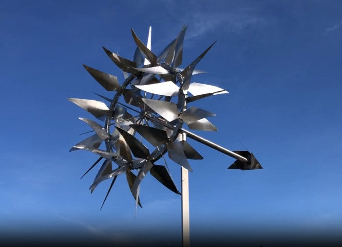 Intricate kinetic sculpture by Martin Jarvis composed of multiple polished stainless steel blades arranged in a radial formation, mounted on a vertical pole with a directional arrow. The piece rotates smoothly with the wind, casting shifting reflections and creating a mesmerising visual effect.