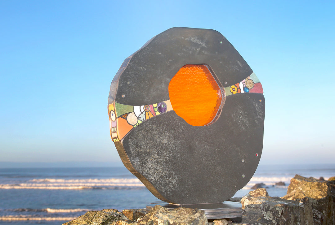 Slate and glass sculpture by Stacey Beaumont titled Sundancer, featuring reclaimed Delabole slate with a glowing amber-orange glass core, inlaid with sea-glass, fossils, ceramics, gemstones, and gold leaf, inspired by coastal light.