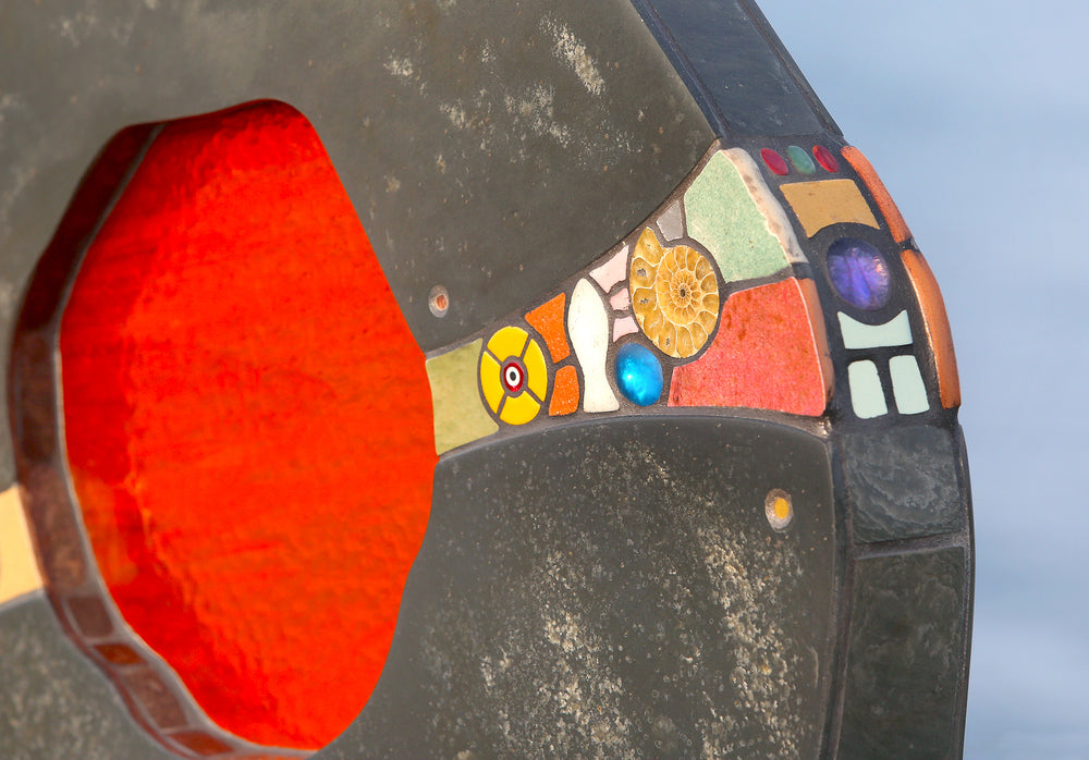 Slate and glass sculpture by Stacey Beaumont titled Sundancer, featuring reclaimed Delabole slate with a glowing amber-orange glass core, inlaid with sea-glass, fossils, ceramics, gemstones, and gold leaf, inspired by coastal light.